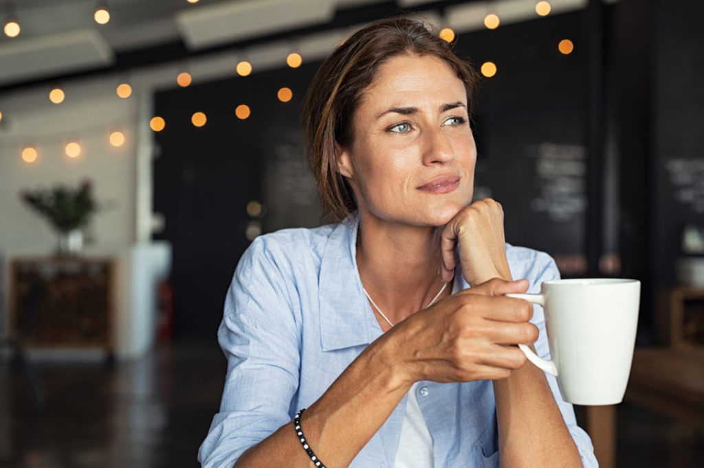 Thoughtful mature woman sitting in cafeteria holding coffee mug while looking away. Middle aged woman drinking tea while thinking. Relaxing and thinking while drinking coffee.